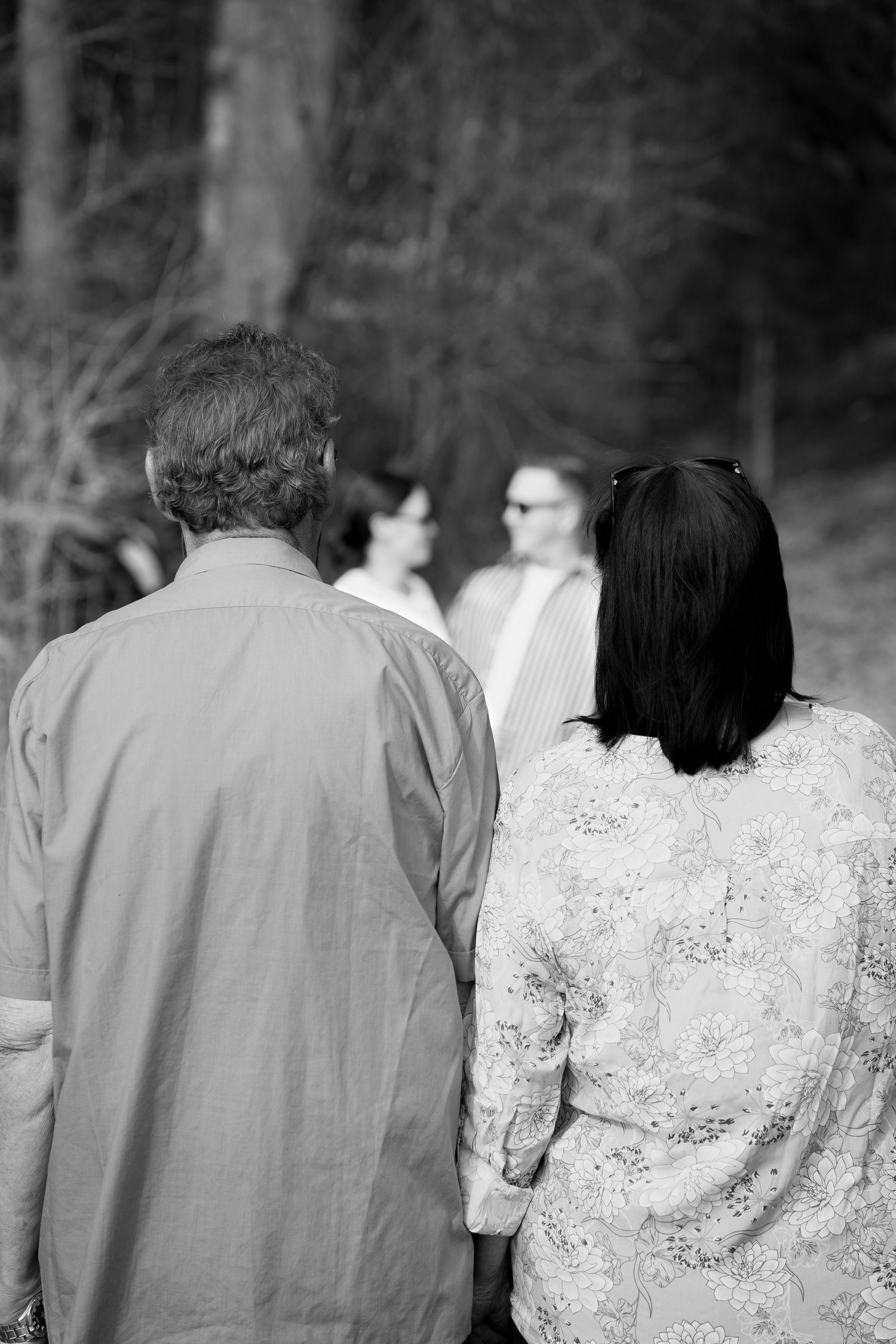 Black and white photo of a couple holding hands from behind, facing others in a wooded outdoor gathering.