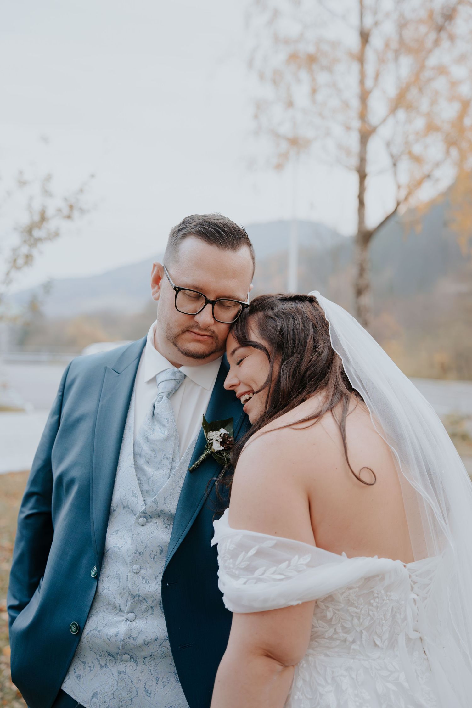 A bride in a white off-shoulder gown and veil leans on her groom in a blue suit outdoors.
