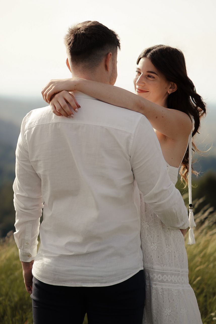 A couple embraces outdoors, woman in white dress smiling over man's shoulder in a sunny field.