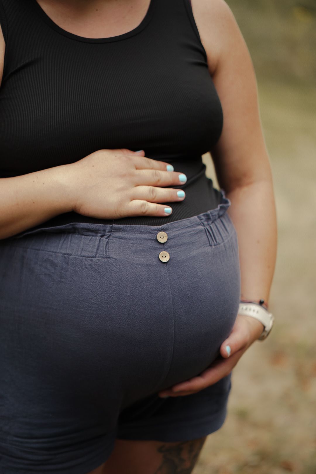 Pregnant woman in black tank top and gray shorts cradling baby bump, showing light blue manicured nails.