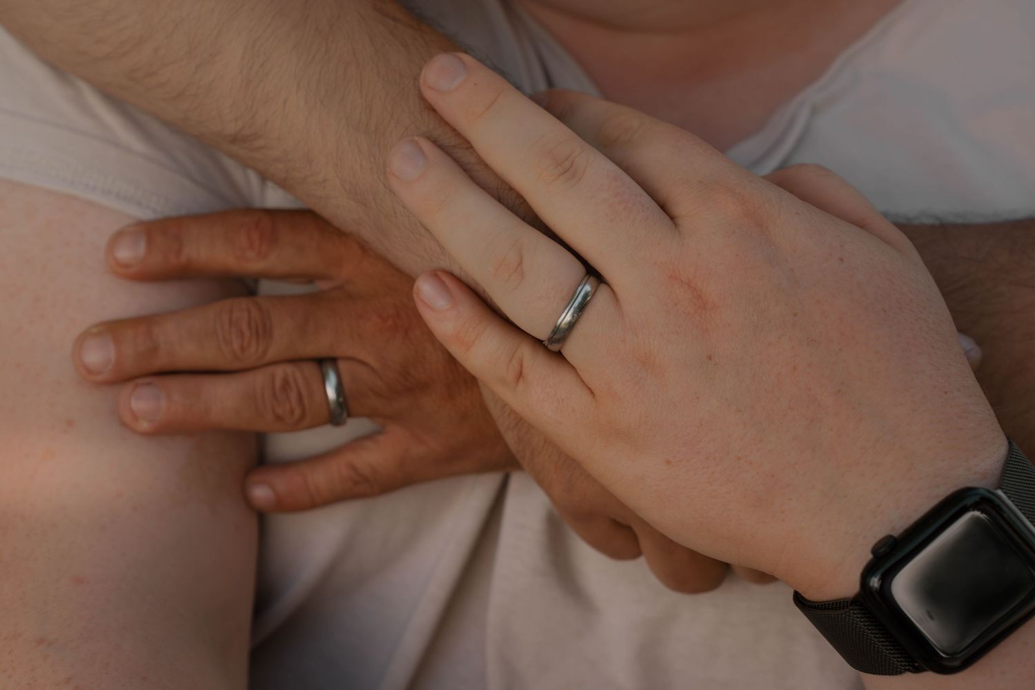 Two hands with matching wedding bands embrace, one wearing a black sports watch, symbolizing marriage and togetherness.
