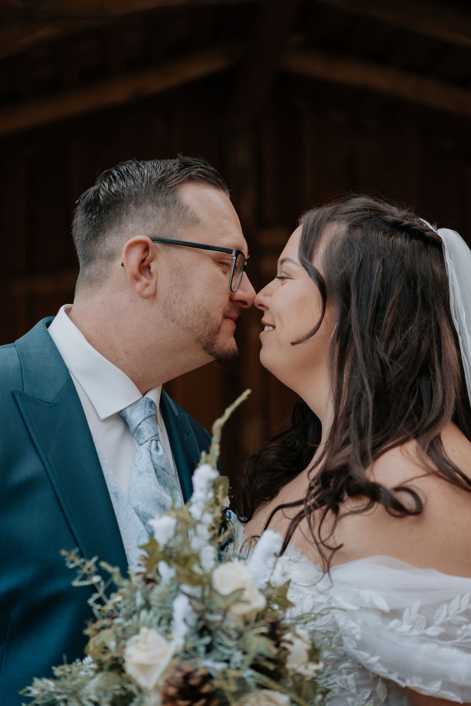 Bride and groom lean in for a kiss, groom in blue suit, bride holding white floral bouquet in rustic barn setting.