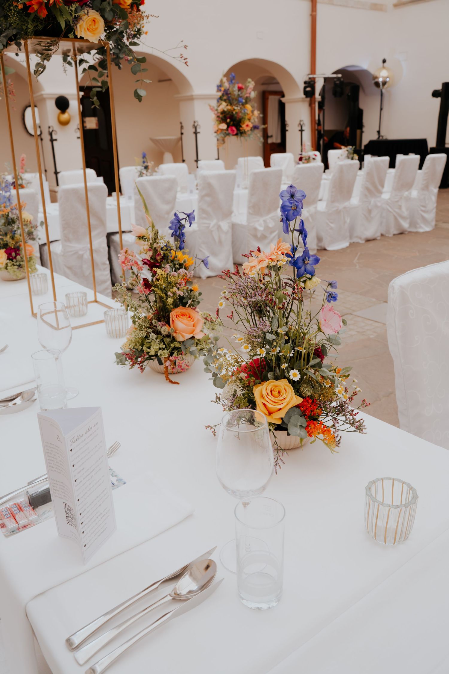 Elegant wedding reception table with colorful wildflower centerpieces, white linens, glassware, and white chair covers in background.