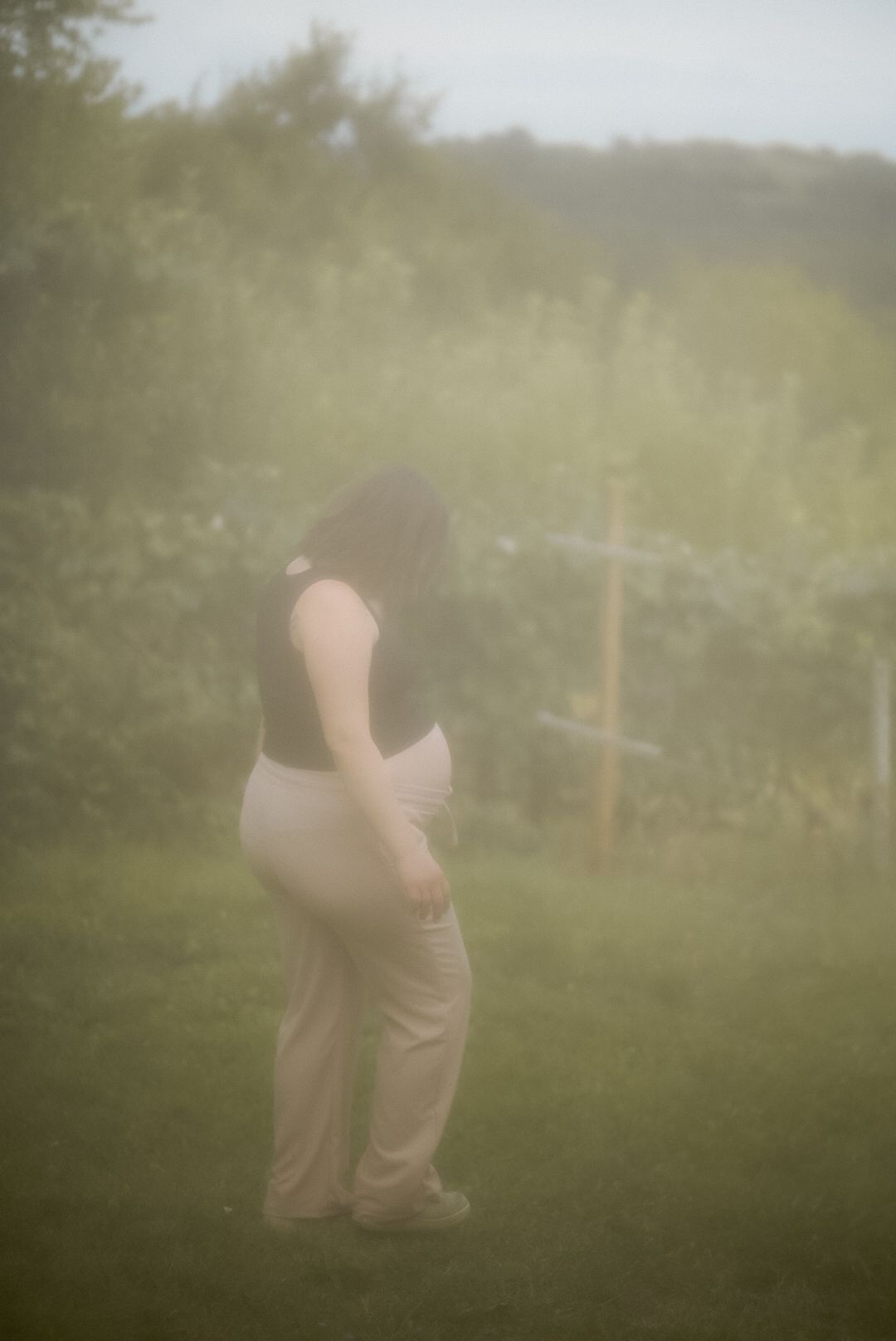 A pregnant woman in a black top and beige pants stands outdoors in a misty garden setting.