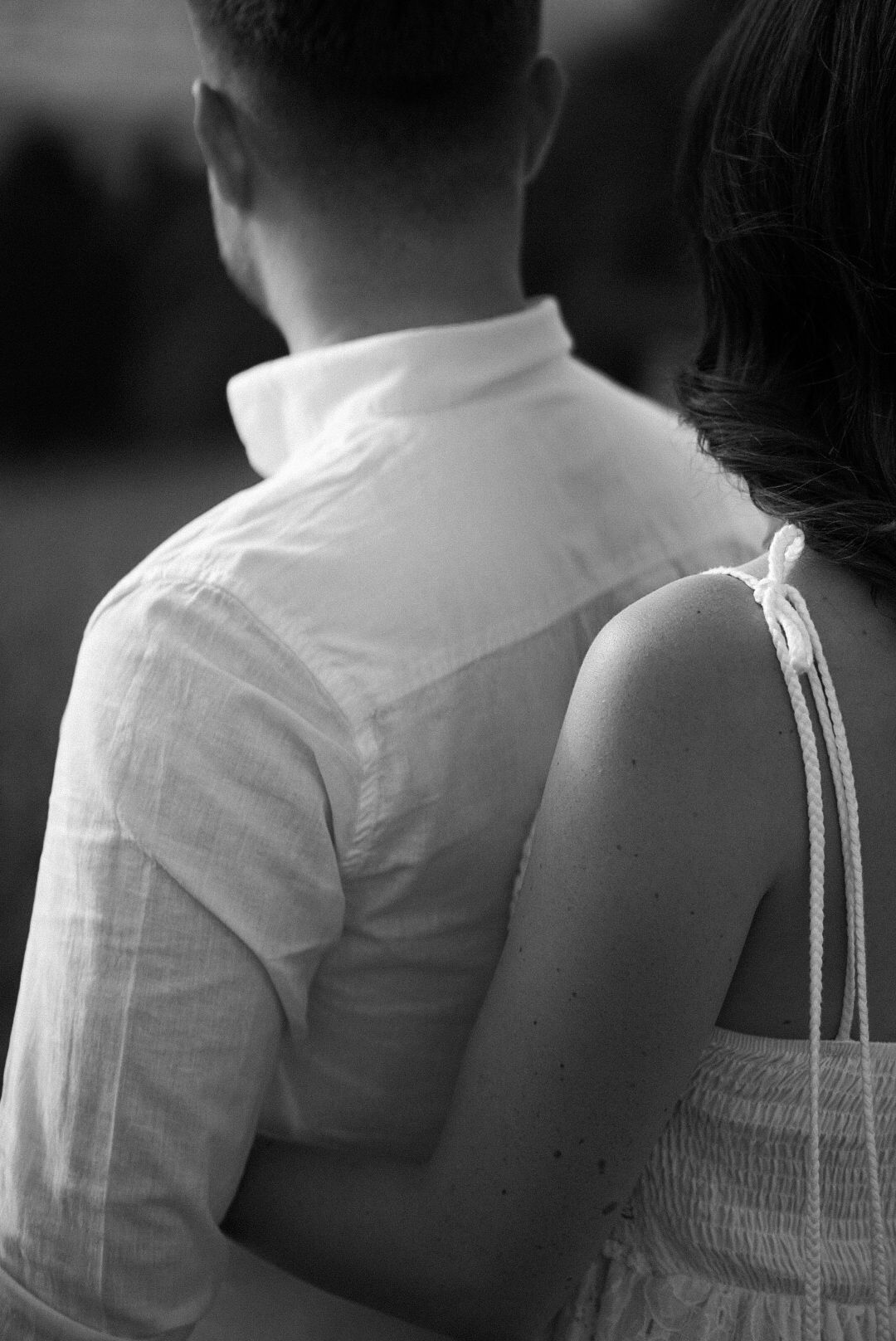 Black and white photo of a couple embracing from behind, man in white linen shirt, woman in strappy dress.