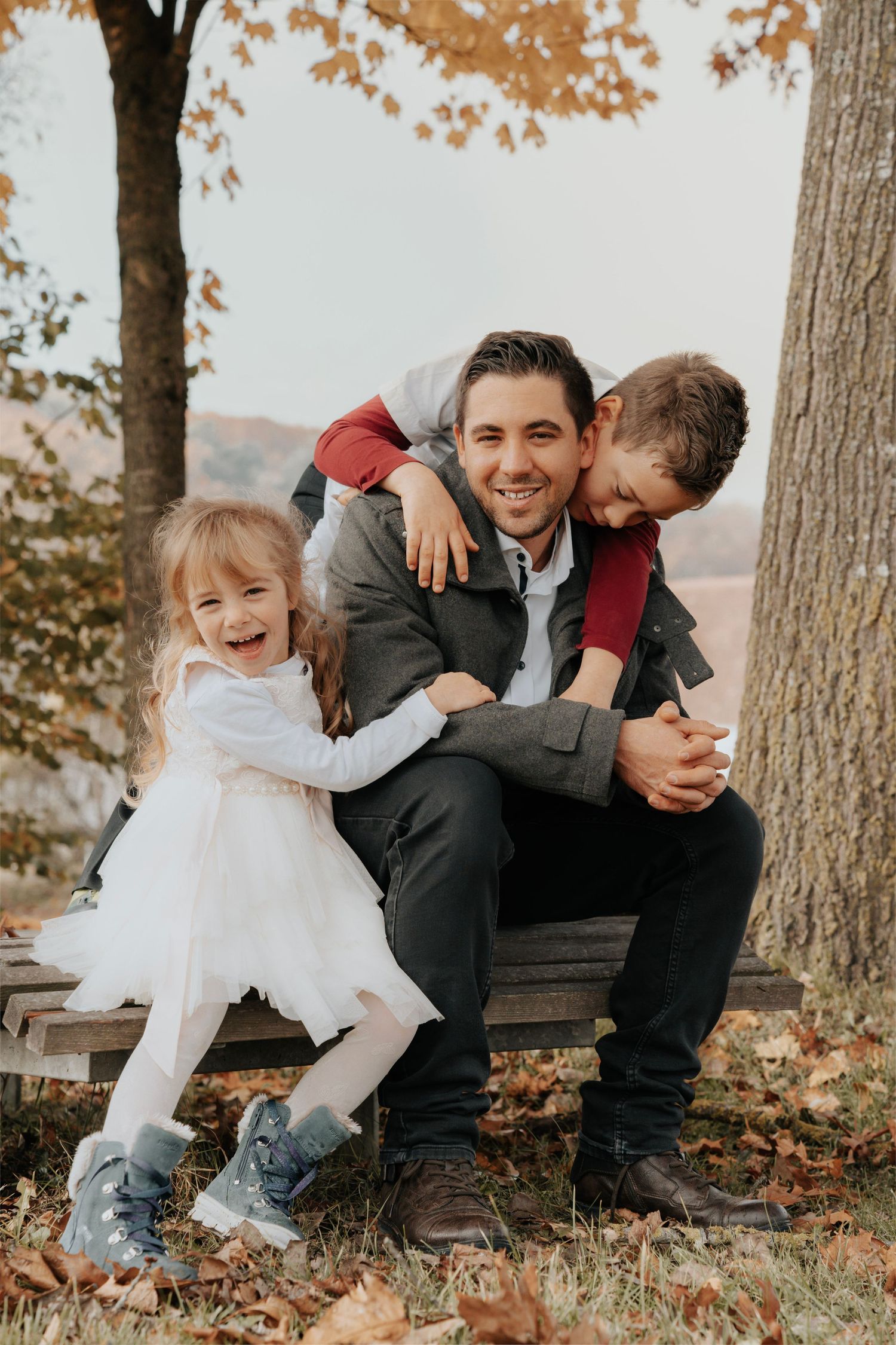 A smiling father sits on a bench with his two young children hugging him outdoors in an autumn park.