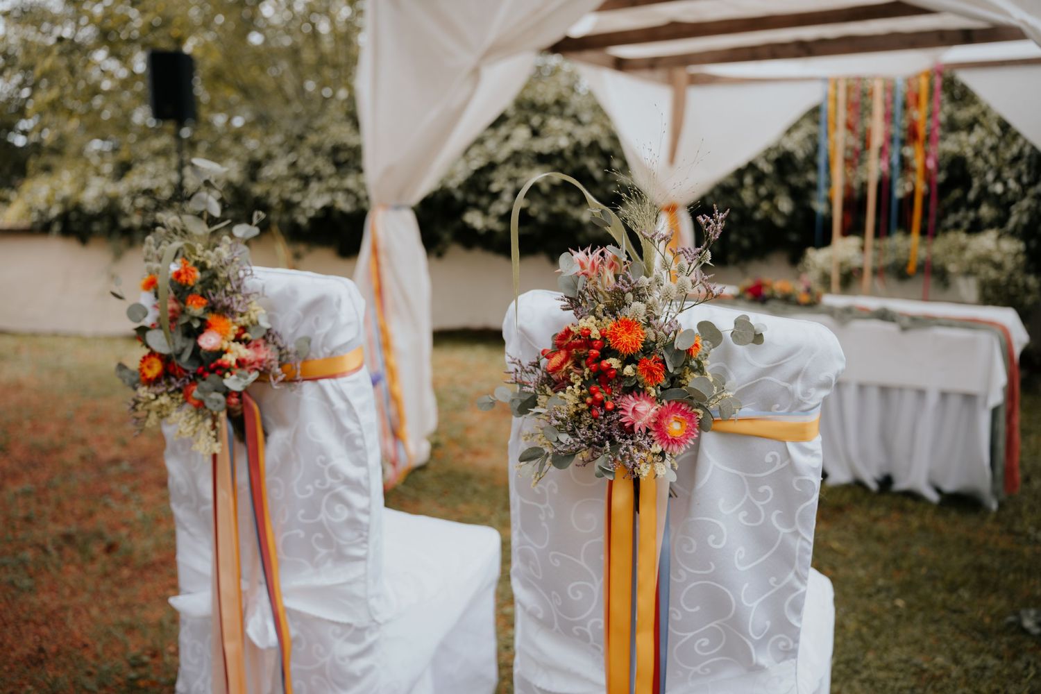White chair covers adorned with colorful floral arrangements and orange ribbons at an outdoor wedding ceremony.