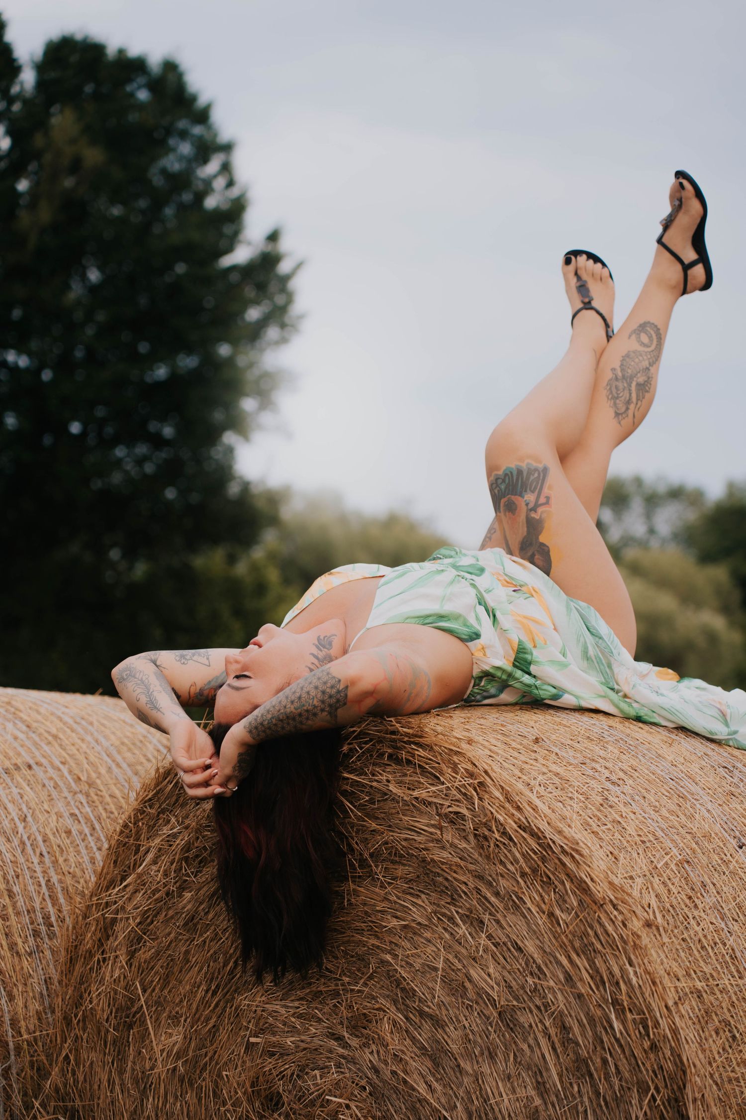 Tattooed woman in floral dress posing on a hay bale with legs raised, wearing black sandals outdoors.