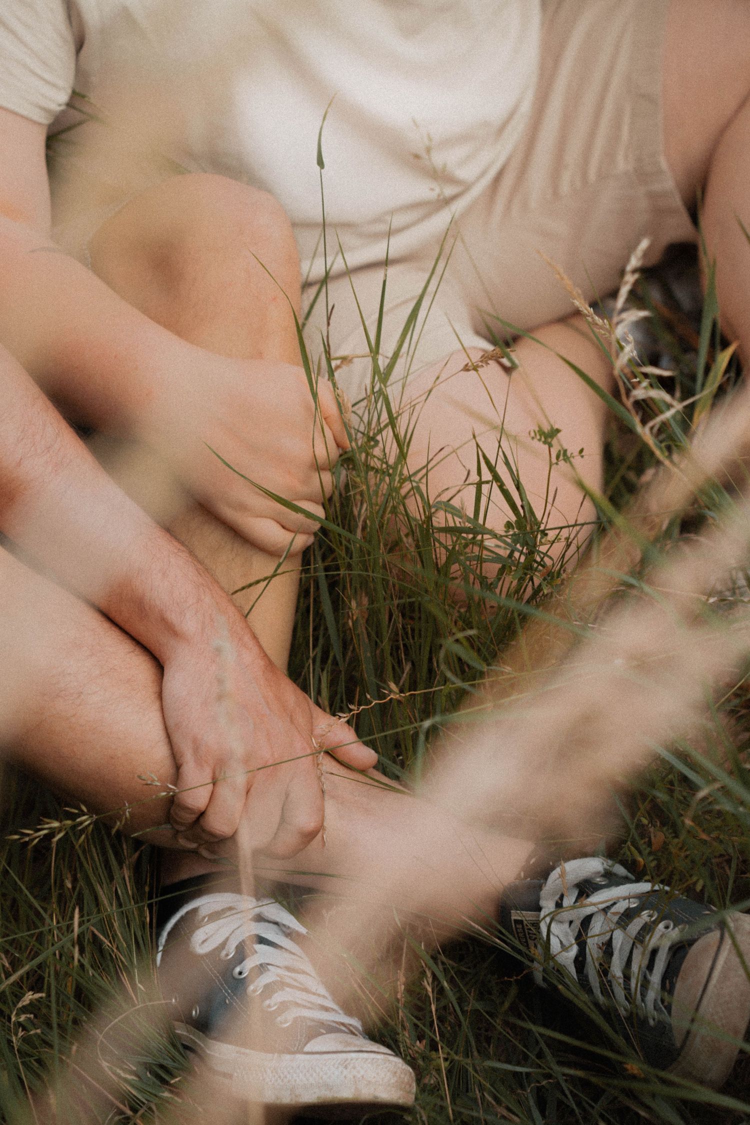 Couple sitting in green grass holding hands, wearing casual white tees and sneakers, romantic outdoor moment.