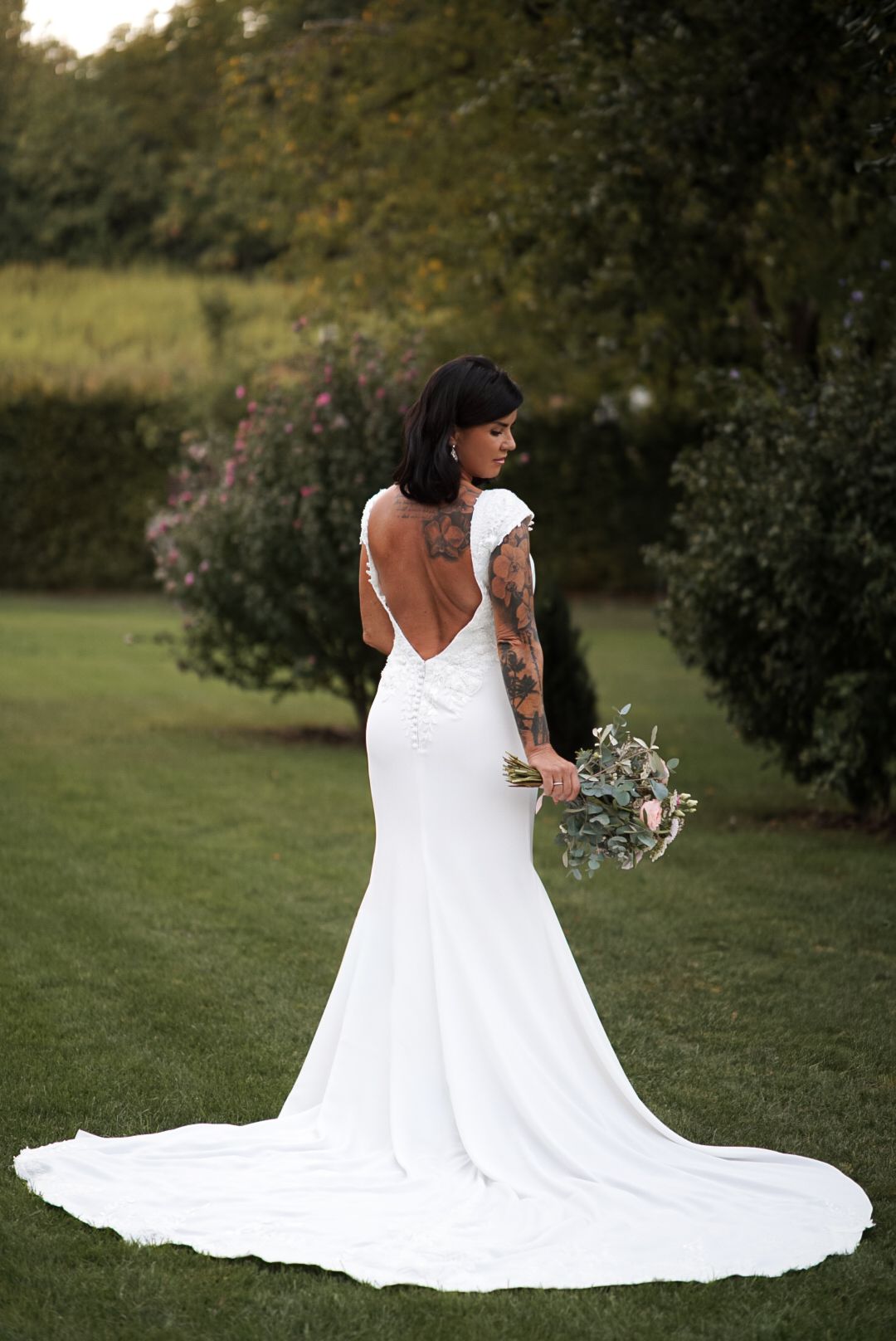 Tattooed bride in backless white mermaid gown with train holds bouquet outdoors in lush garden setting.