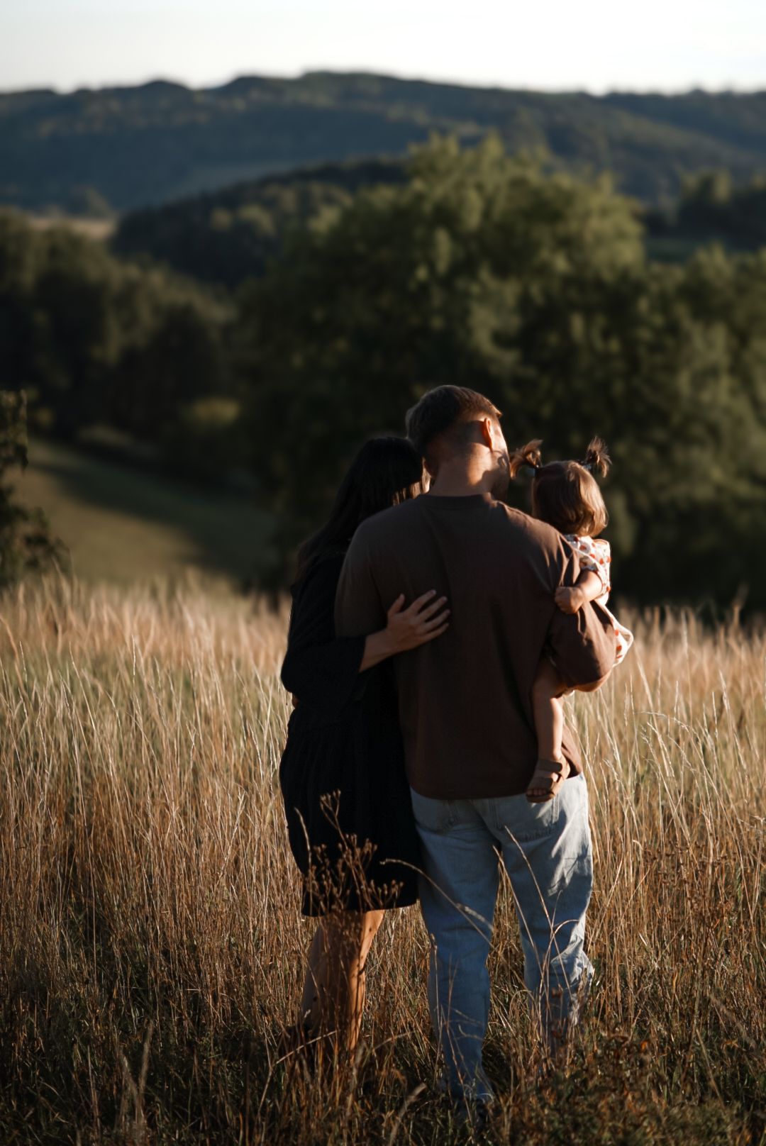A family embraces in a golden field at sunset with rolling hills and trees in the background.