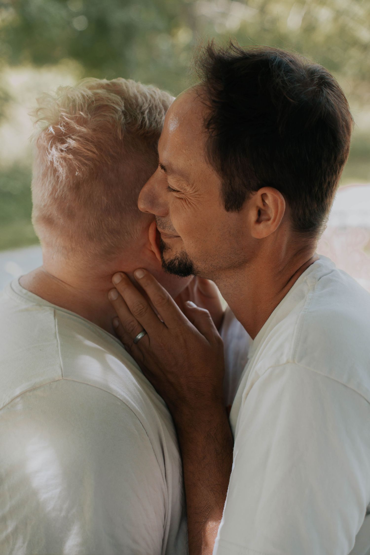 Two men sharing an intimate moment, one gently cupping the other's face, both wearing white shirts outdoors.