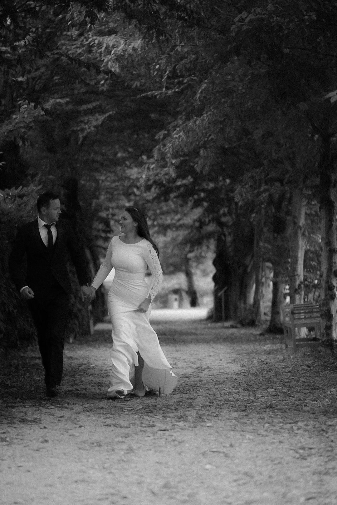 Black and white photo of a bride and groom holding hands, walking along a tree-lined path on their wedding day.