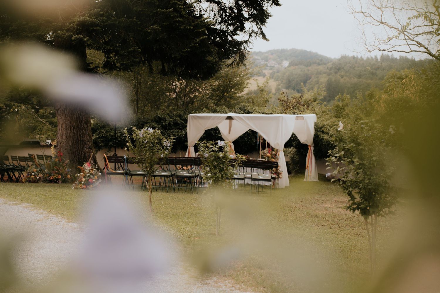 Outdoor wedding ceremony setup with draped canopy altar, rustic wooden benches, and lush green hillside backdrop.