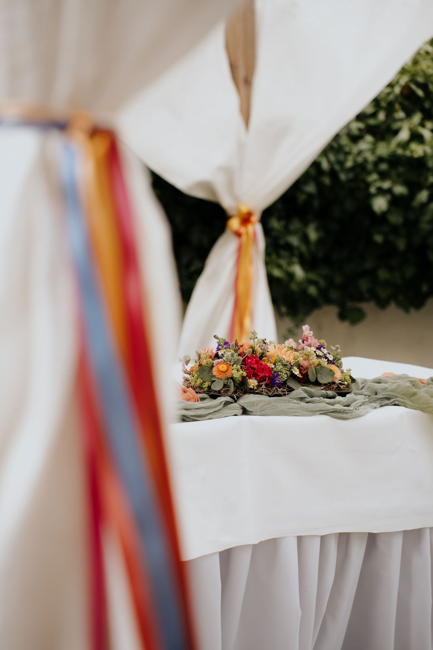 Elegant wedding ceremony table with colorful ribbon drapes and vibrant floral arrangement on white linen outdoors.