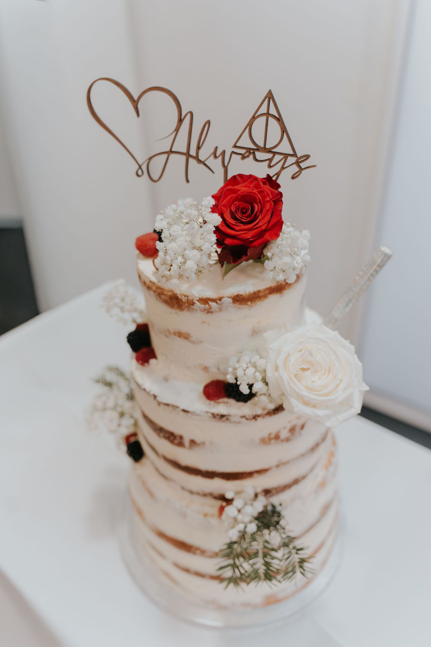 Three-tier naked wedding cake with red roses, berries, and Harry Potter Always cake topper.