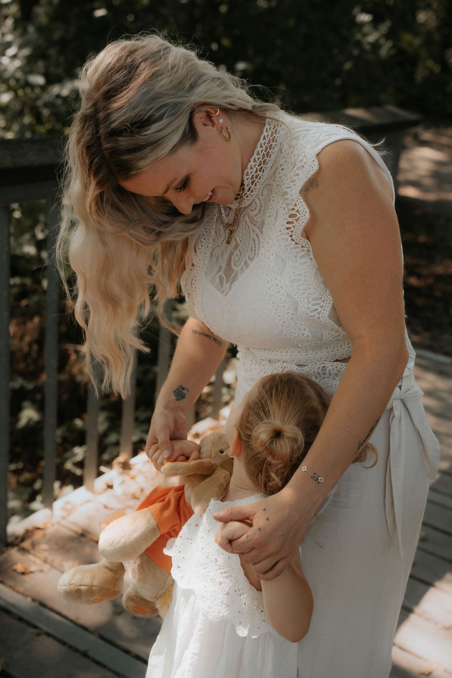 Bride in white lace dress smiling at young flower girl holding teddy bears on wooden deck.