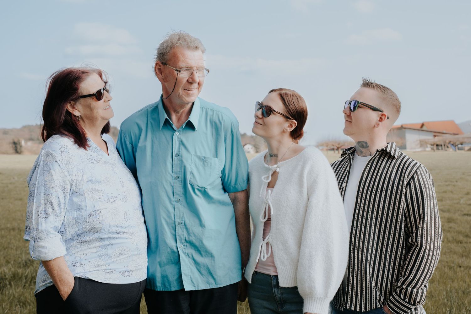 Four people wearing sunglasses smile and pose together outdoors in a grassy field on a sunny day.