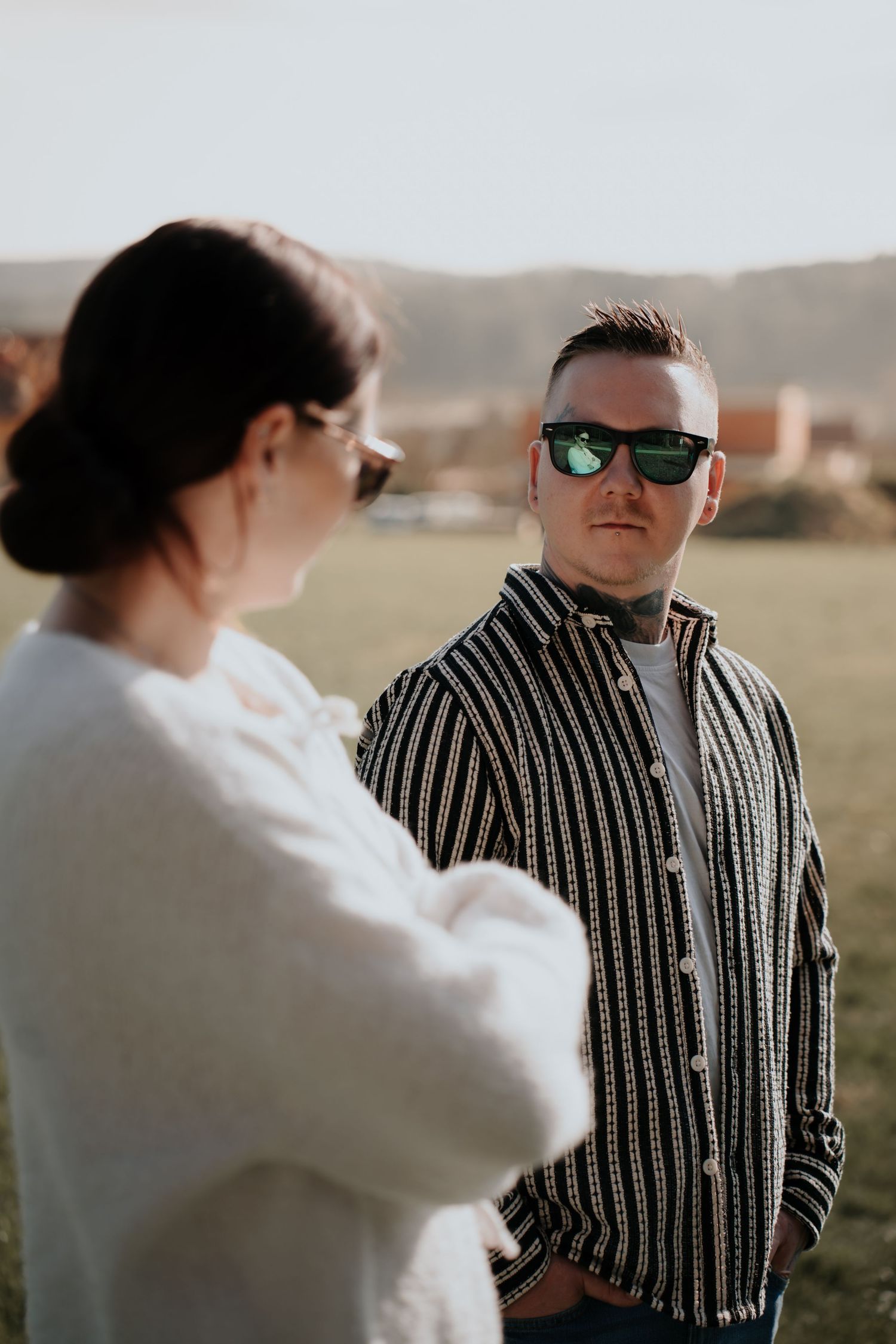 Man in striped shirt and sunglasses stands outdoors facing a woman in white, with green hills in the background.