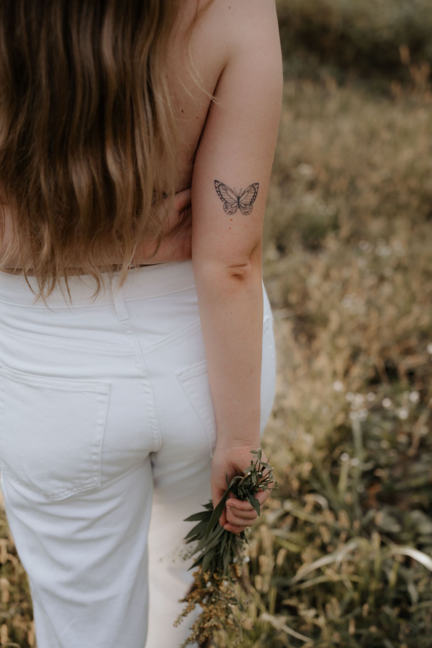 Woman in white jeans with butterfly tattoo on arm holding wildflowers in a field.