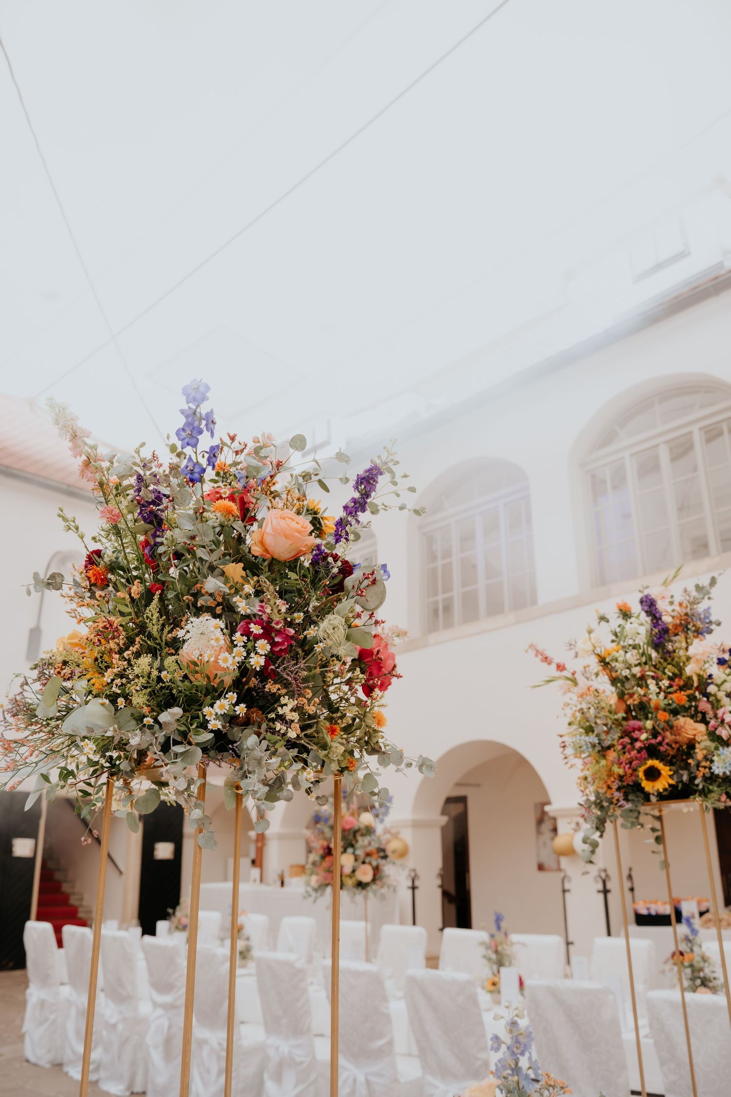 Tall gold stands with colorful wildflower arrangements decorate a white venue with chair covers for a wedding ceremony.