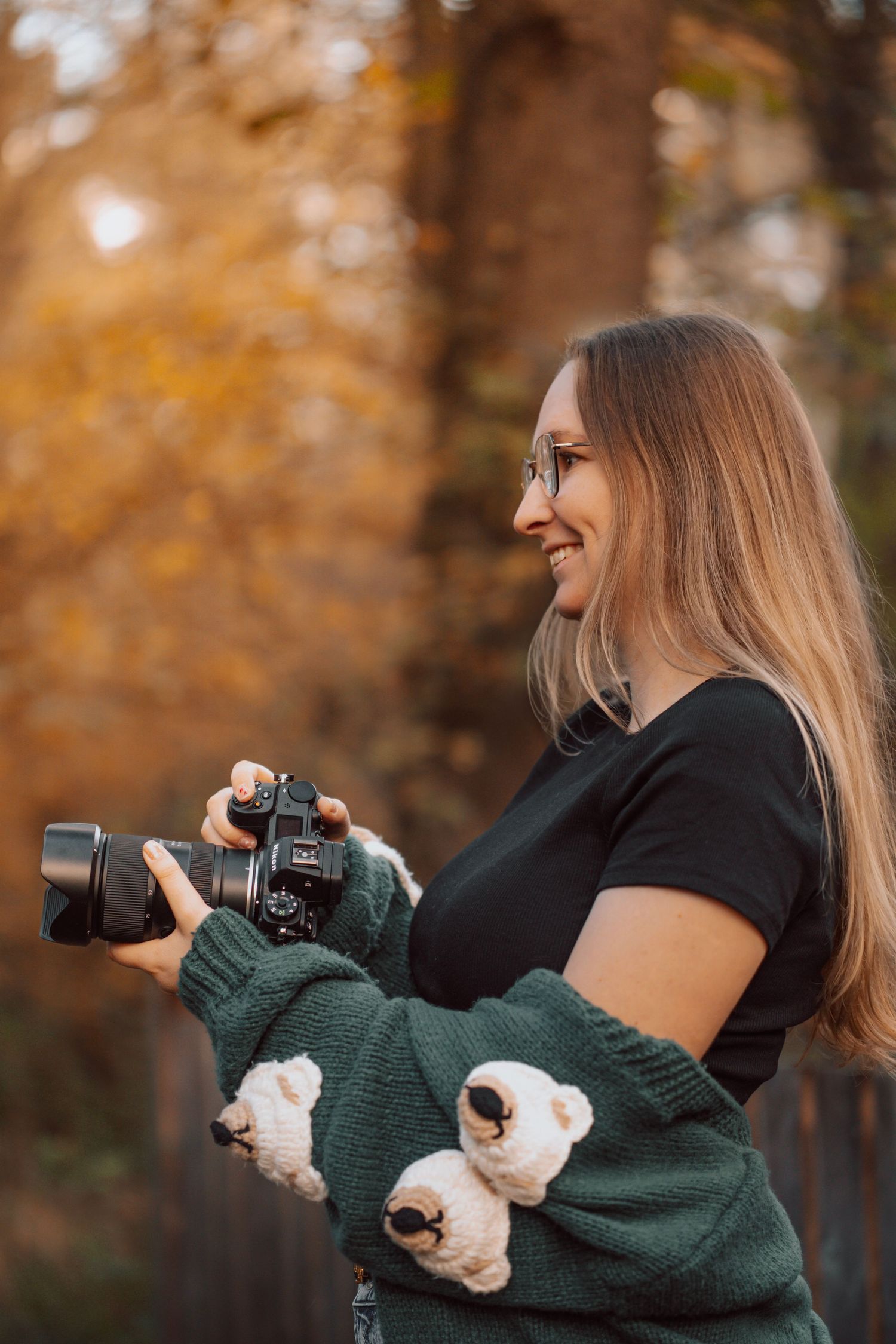 Smiling woman with glasses holding a DSLR camera outdoors in autumn forest, wearing green sweater with bear design.