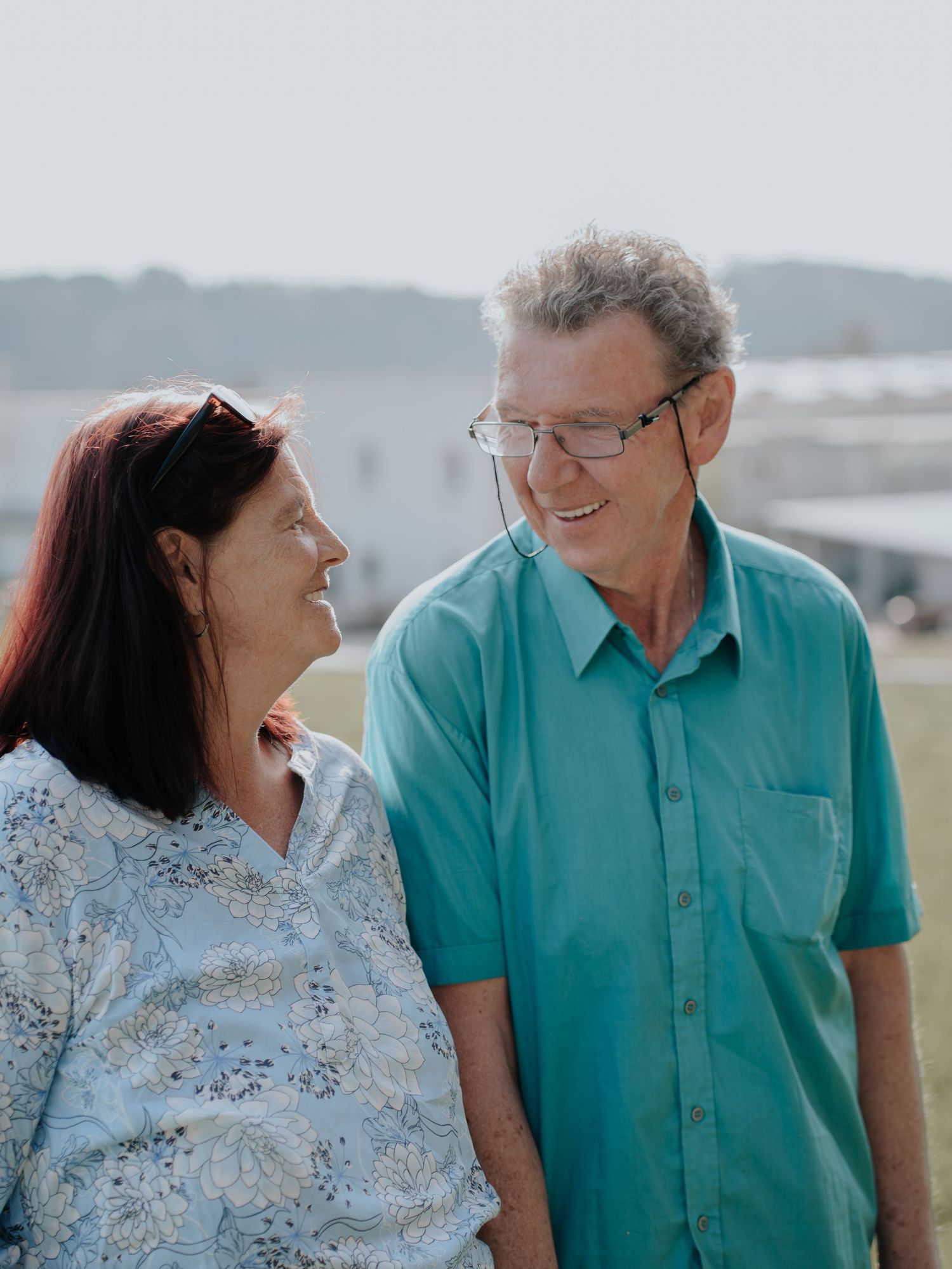 An older couple smiling at each other outdoors on a sunny day, woman with red hair and man wearing glasses and teal shirt.