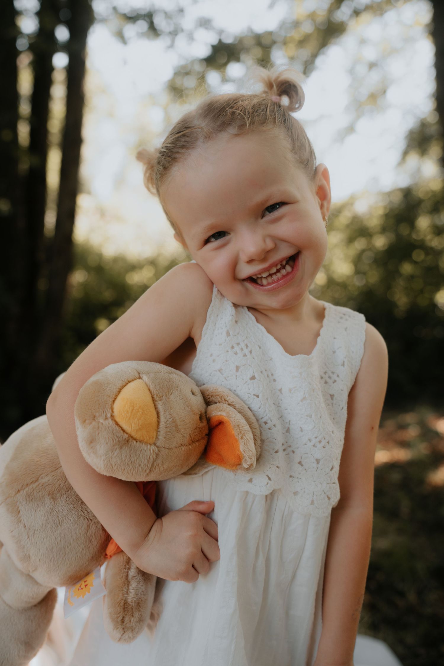 A joyful toddler girl in a white dress hugs a stuffed animal toy while smiling brightly outdoors.