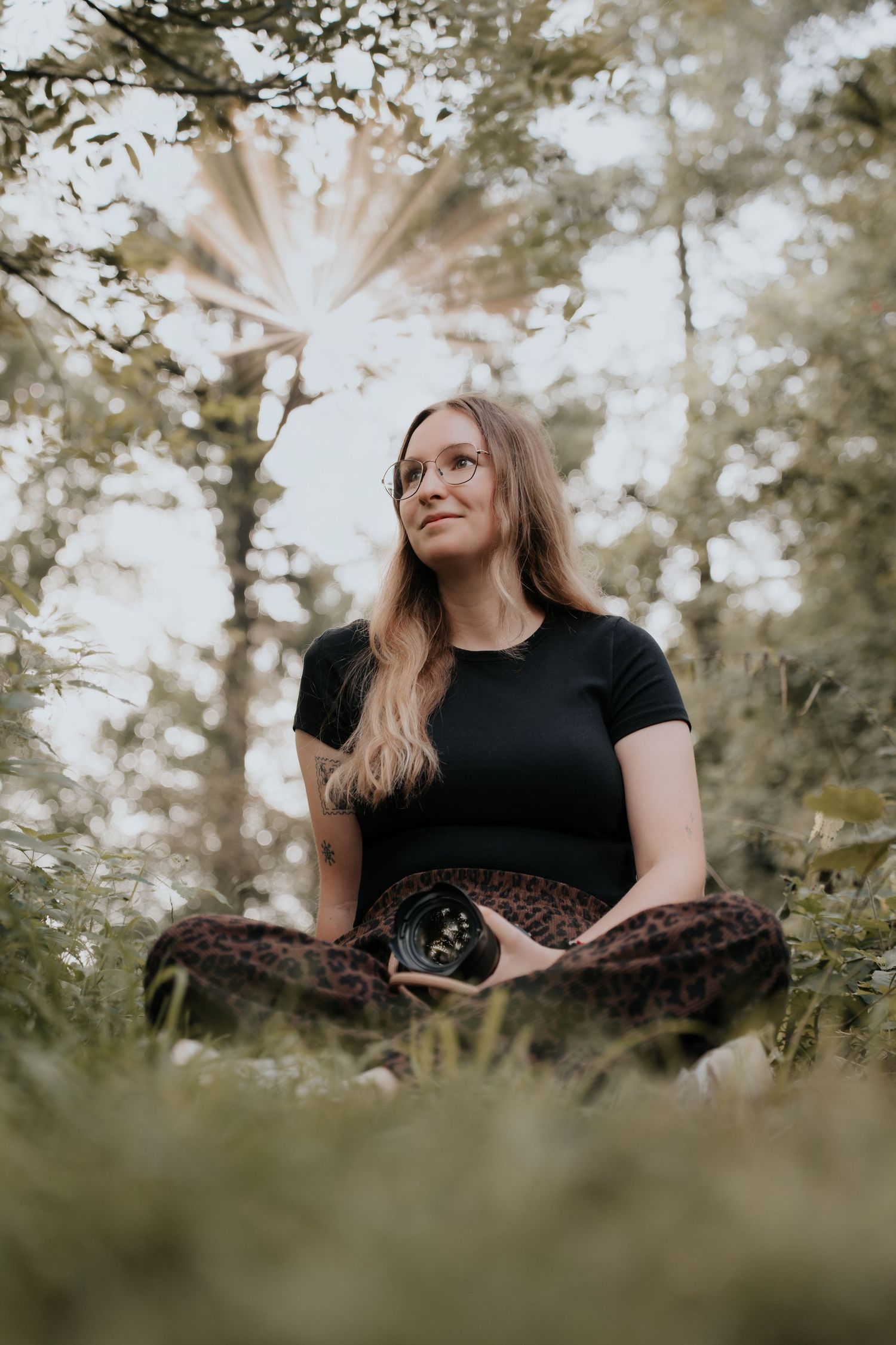 Woman in black tee and leopard pants meditating cross-legged in lush green forest with sunlight filtering through trees.