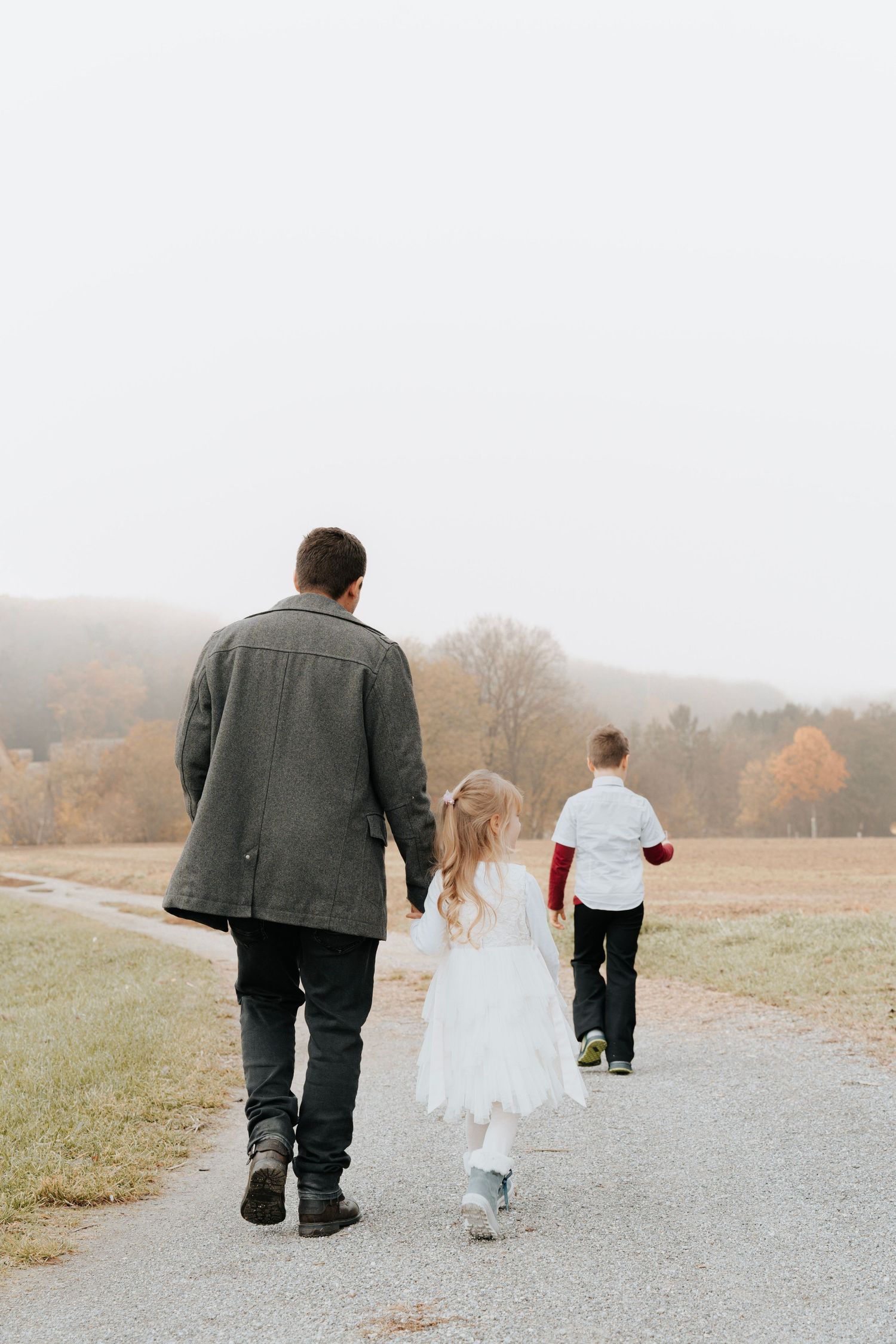 A father walks hand-in-hand with his young daughter and son along a misty rural path in fall.
