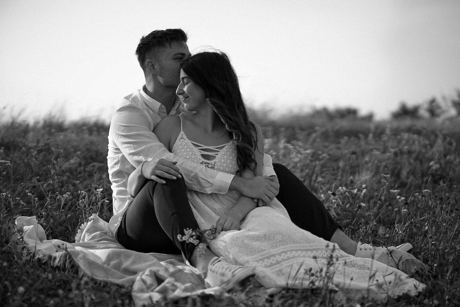Black and white photo of a romantic couple sitting together in a field, the man embracing the woman from behind.