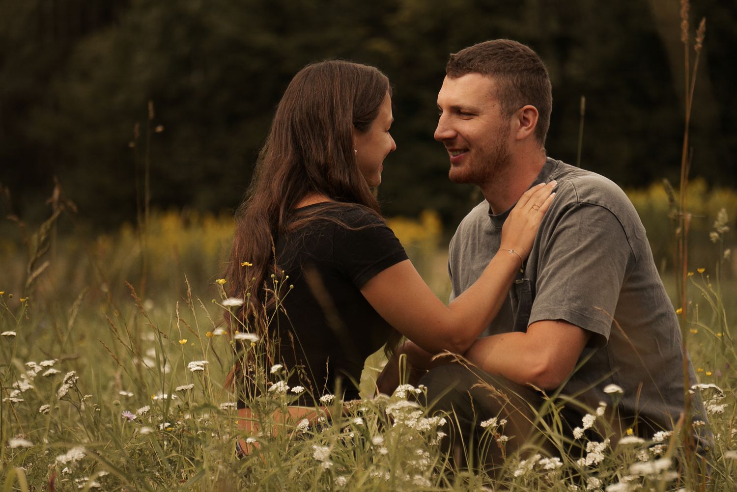 A couple sits romantically in a wildflower meadow, gazing at each other with smiles in a lush green field.