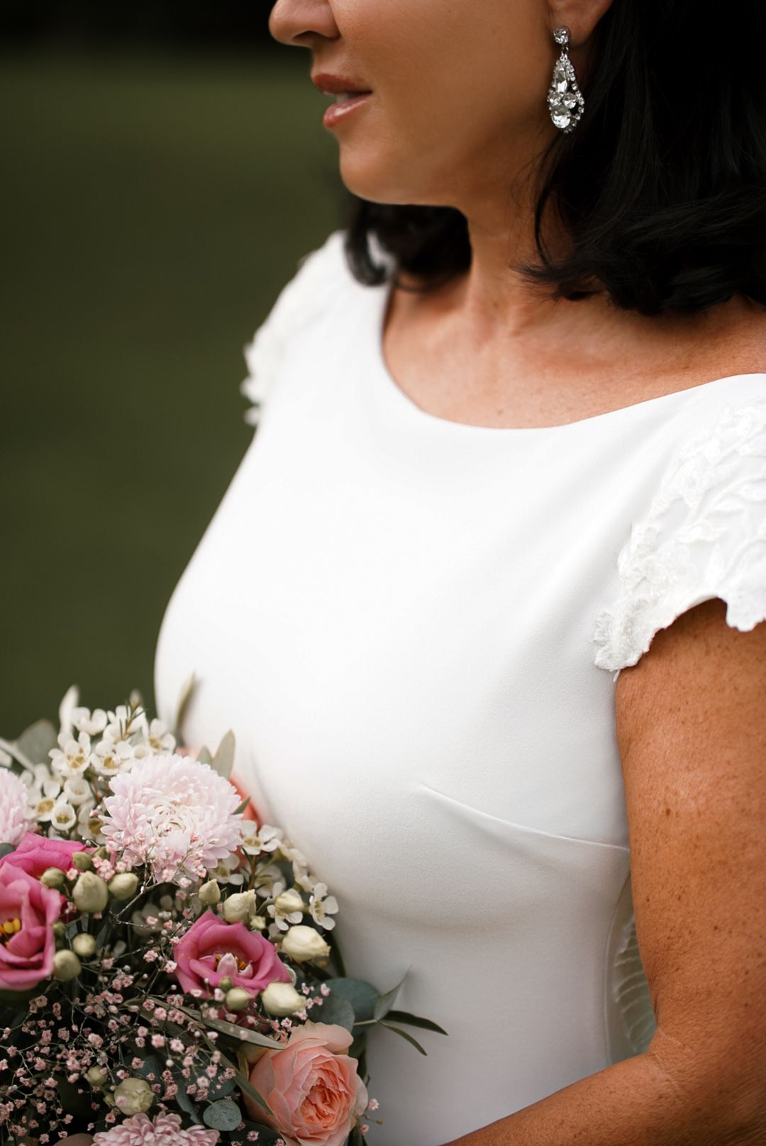 Bride in white wedding dress holding pink floral bouquet, wearing crystal drop earrings, close-up portrait shot.