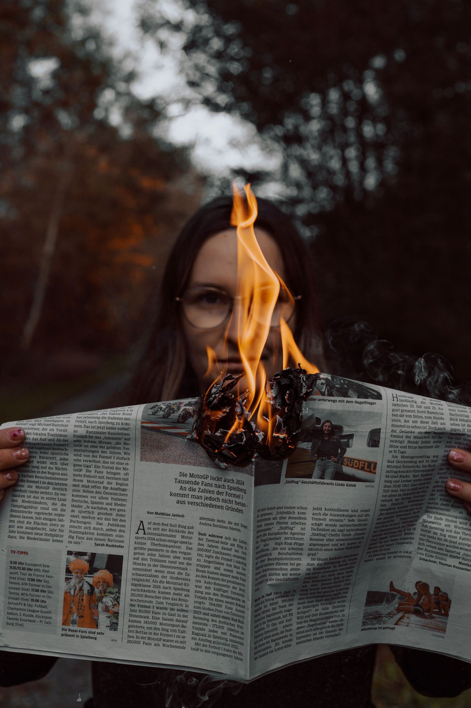 A woman holds a burning newspaper with flames rising from its center outdoors.