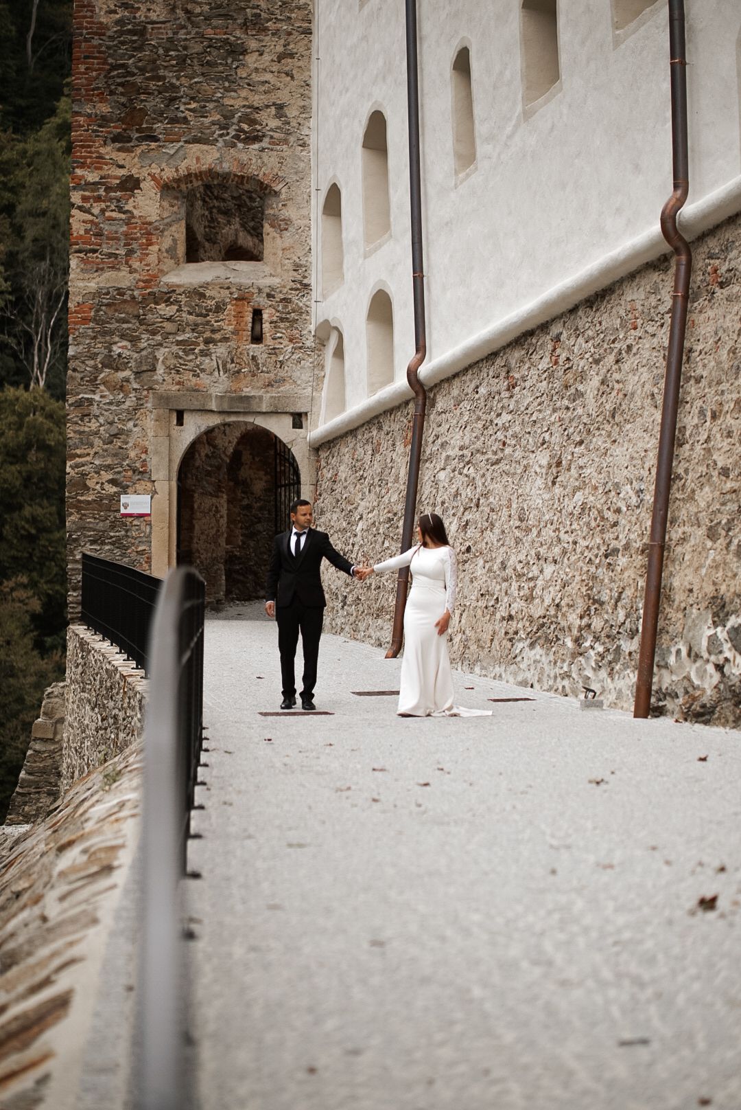 Bride and groom hold hands walking along a stone castle wall pathway during their wedding day photoshoot.