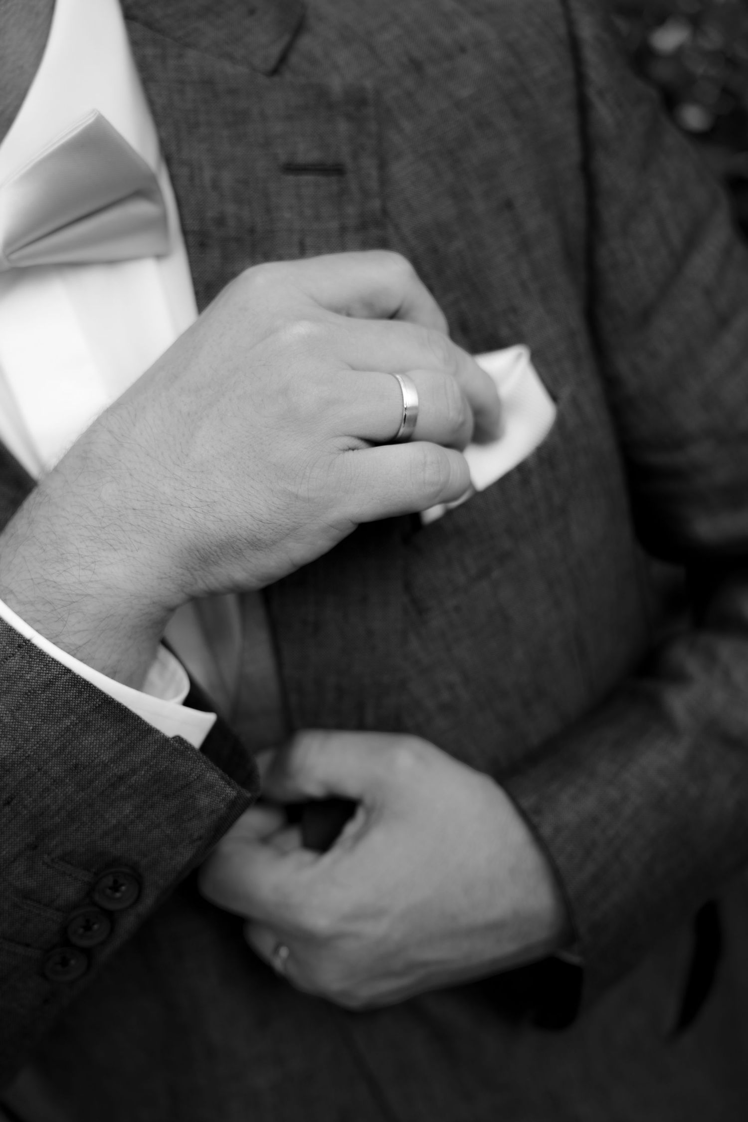 Black and white photo of a groom adjusting his pocket square, wearing a wedding ring and tailored suit.