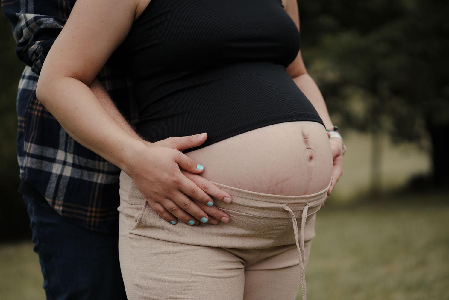 Pregnant woman in black top and beige pants holds baby bump outdoors with partner beside her.
