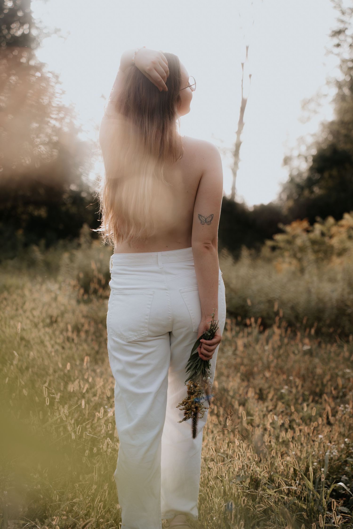 Woman with long hair and butterfly tattoo stands in golden field holding wildflowers, wearing white pants, back to camera.