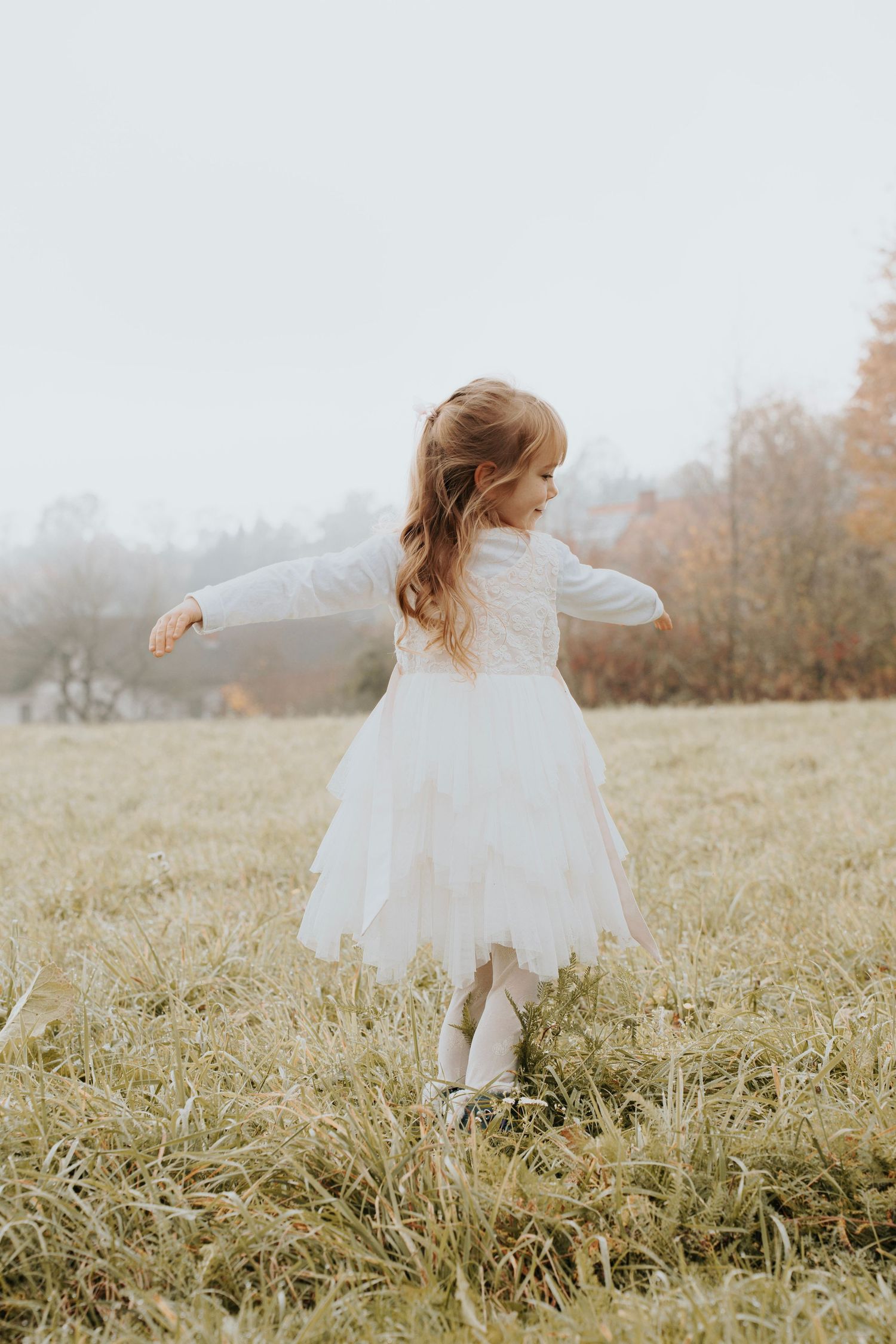 Young girl in white tulle dress twirling in an autumn meadow, arms outstretched, with misty trees in background.