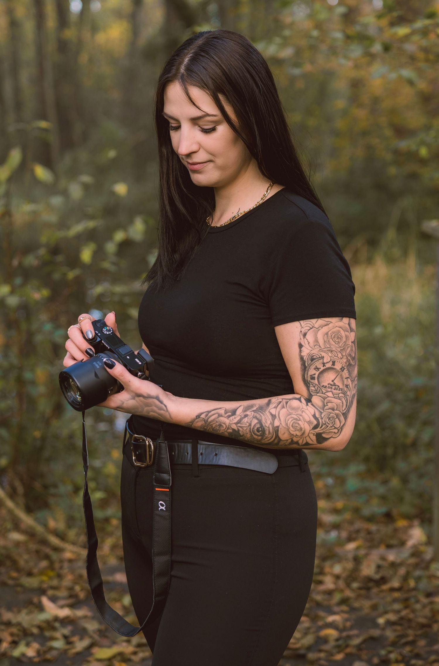 A photographer with tattoos wearing black holds a professional camera in a wooded outdoor setting.
