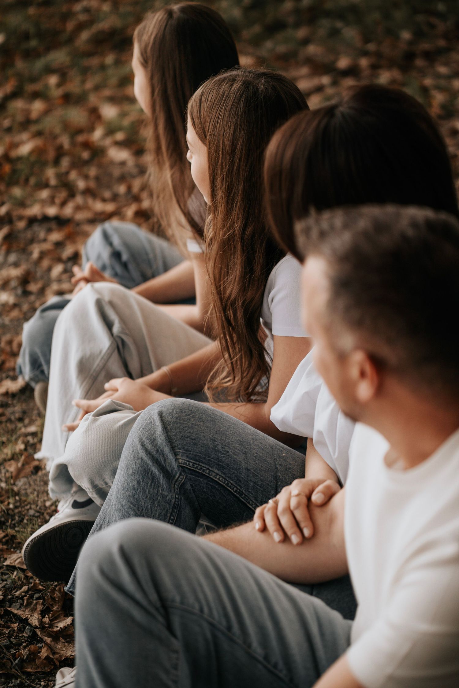 Family sitting together on autumn leaves in a park, wearing white shirts and jeans, enjoying a peaceful outdoor moment.