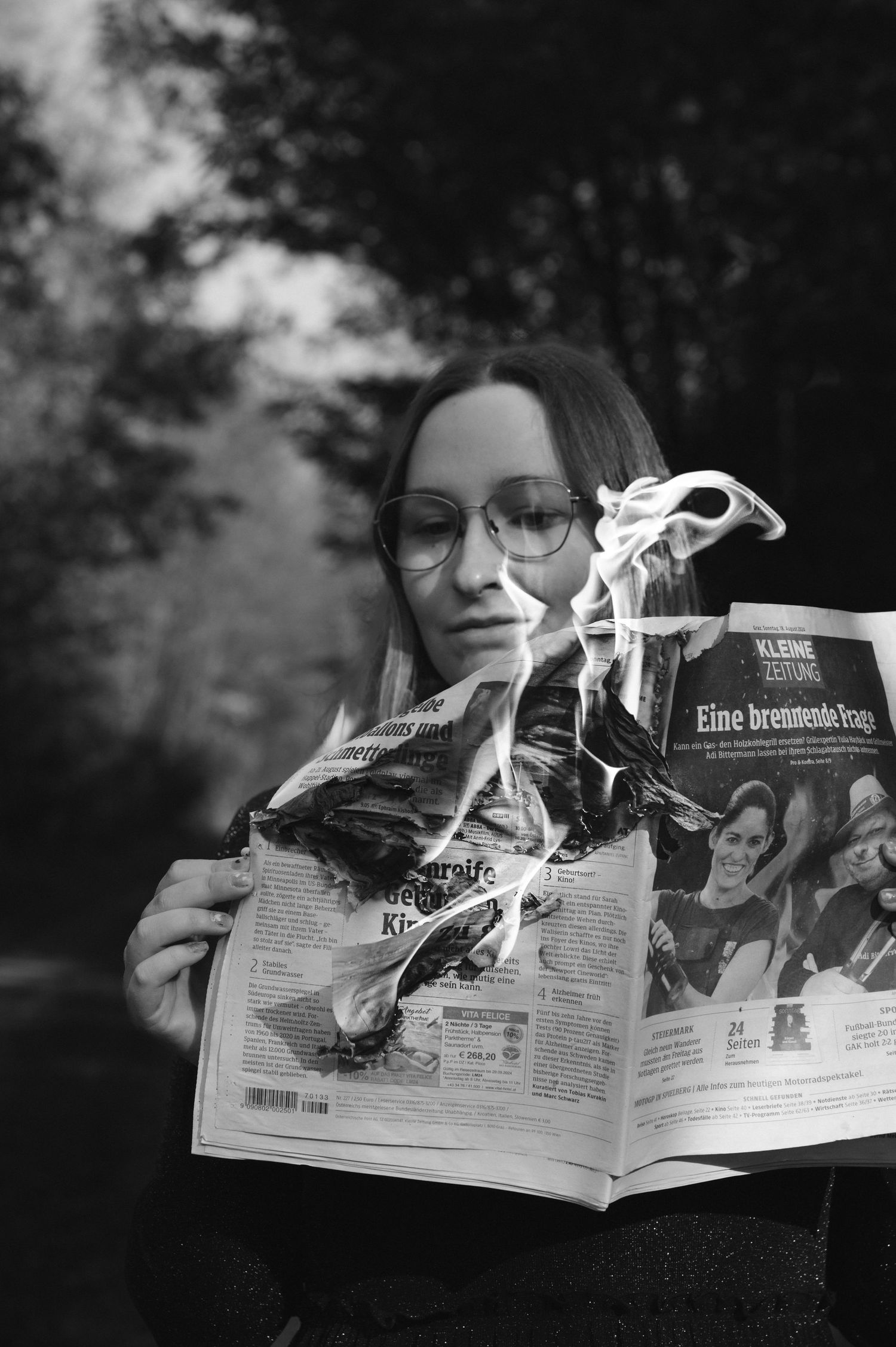 Black and white photo of a woman with glasses reading a burning newspaper outdoors.