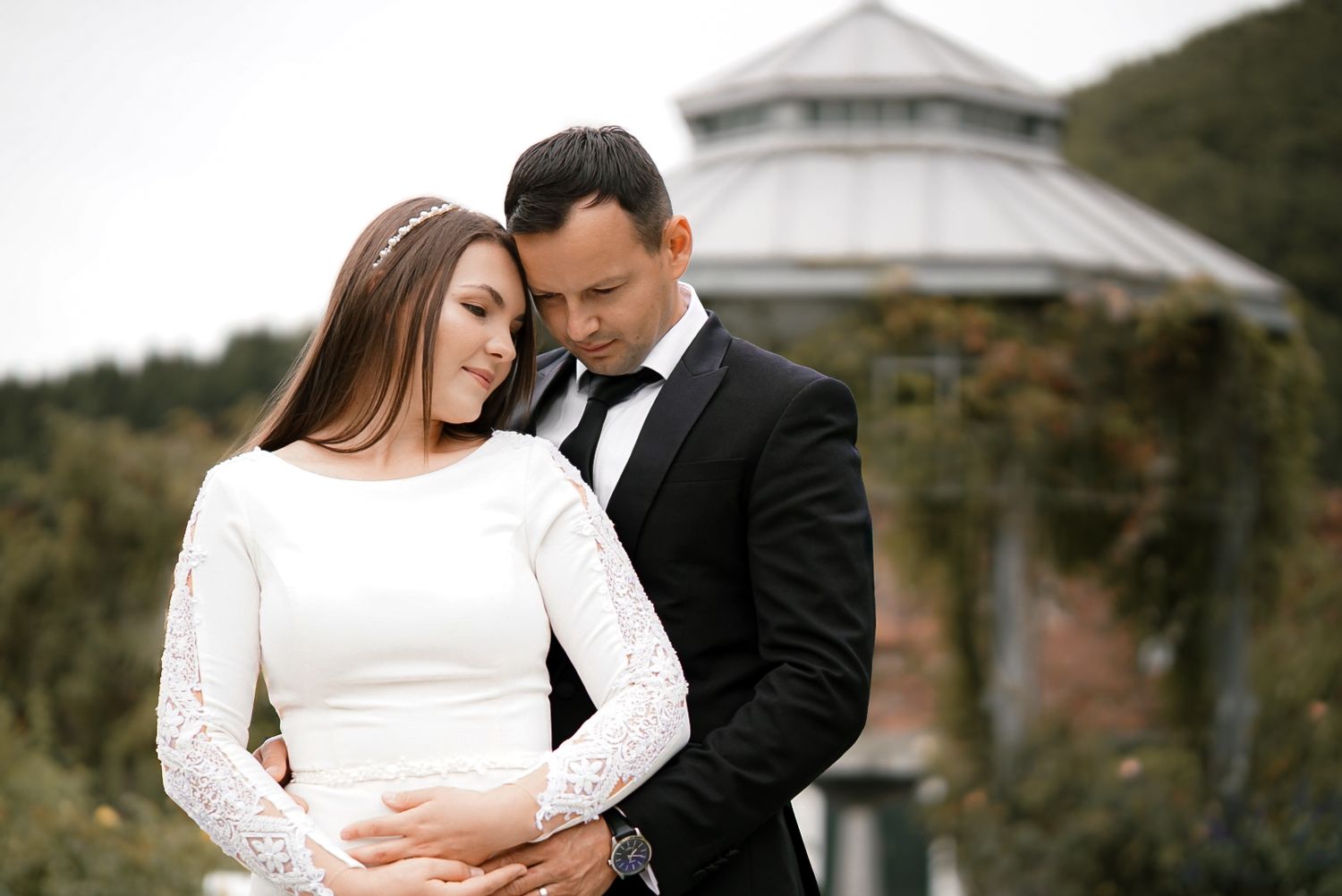 Elegant couple in wedding attire embracing outdoors, bride in white lace gown and groom in black tuxedo.
