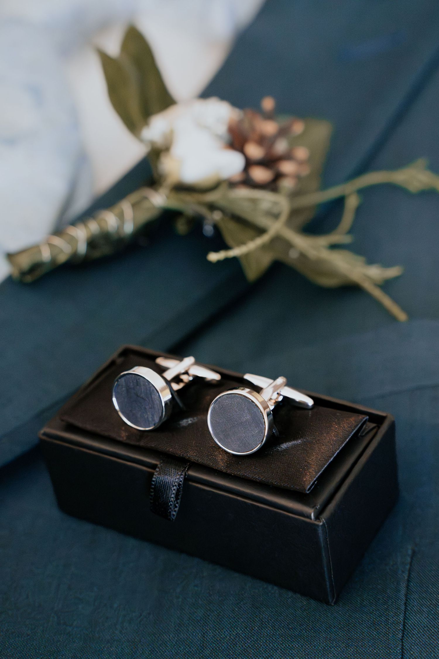 Silver round cufflinks in a black gift box beside a floral boutonniere on a dark teal suit jacket.
