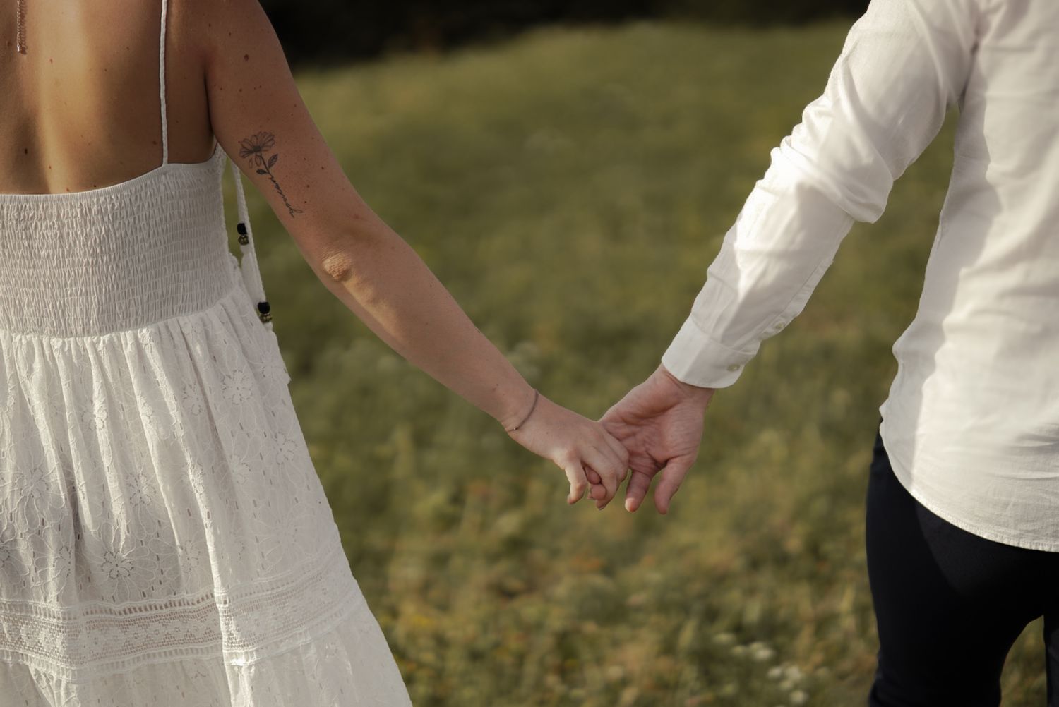 Couple holding hands in a field, woman in white sundress and man in white shirt, romantic outdoor setting.