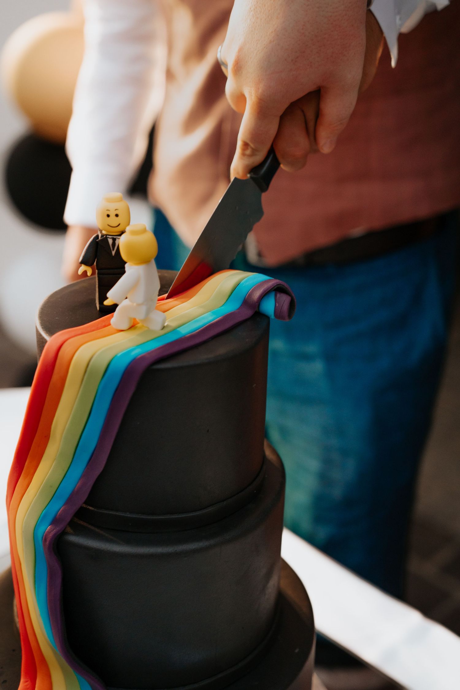 Black tiered wedding cake with rainbow fondant drape and LEGO bride and groom toppers being cut with a knife.