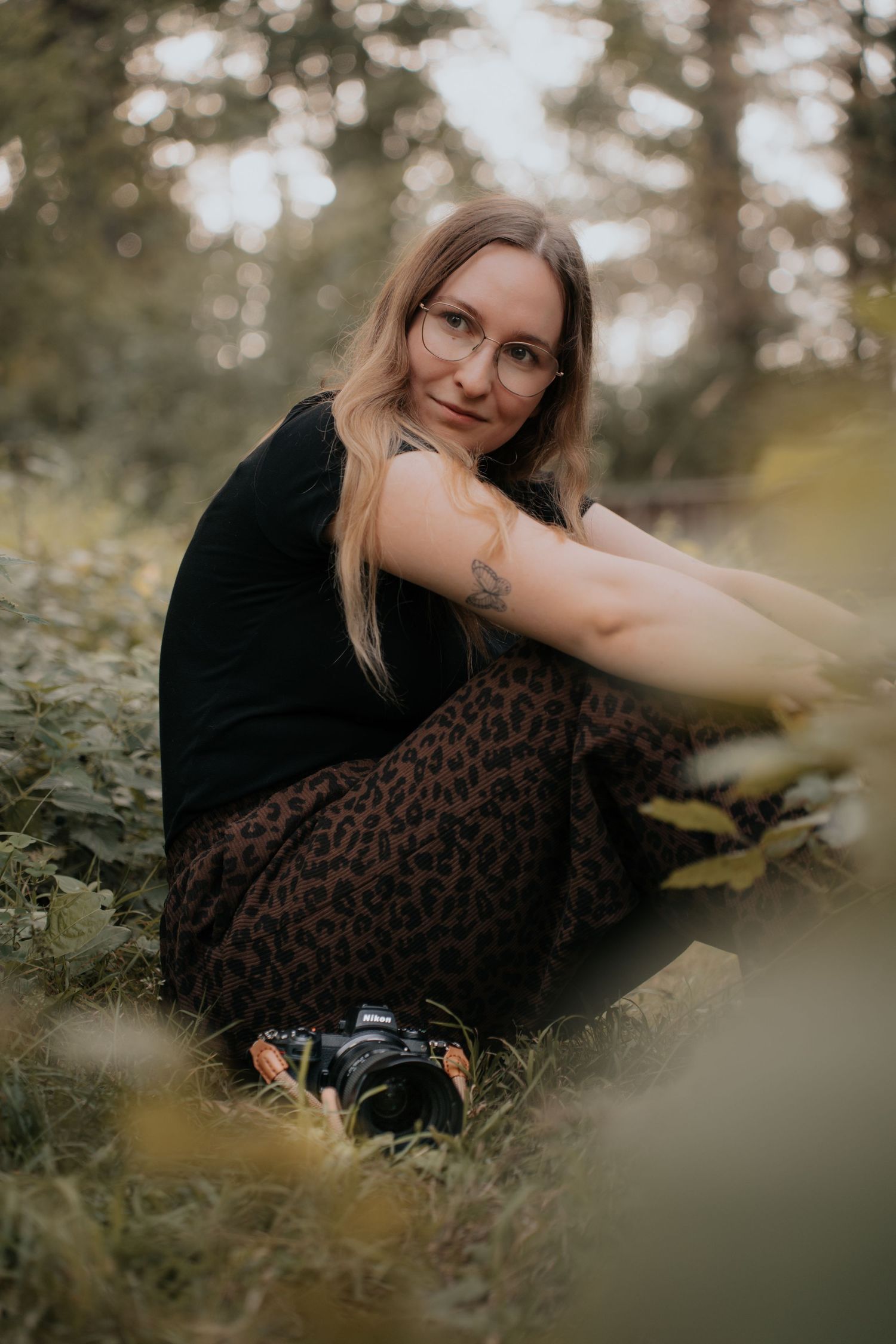 Young woman with glasses sitting in forest grass, wearing black top and leopard skirt, holding a camera.