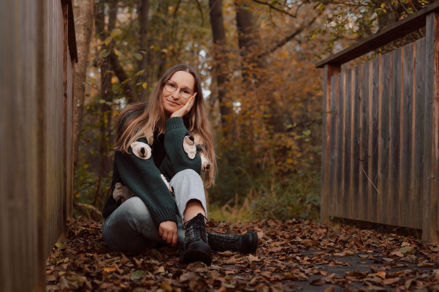 Young woman sitting on autumn leaves outdoors, wearing a green sweater and black boots, smiling beside a wooden fence.