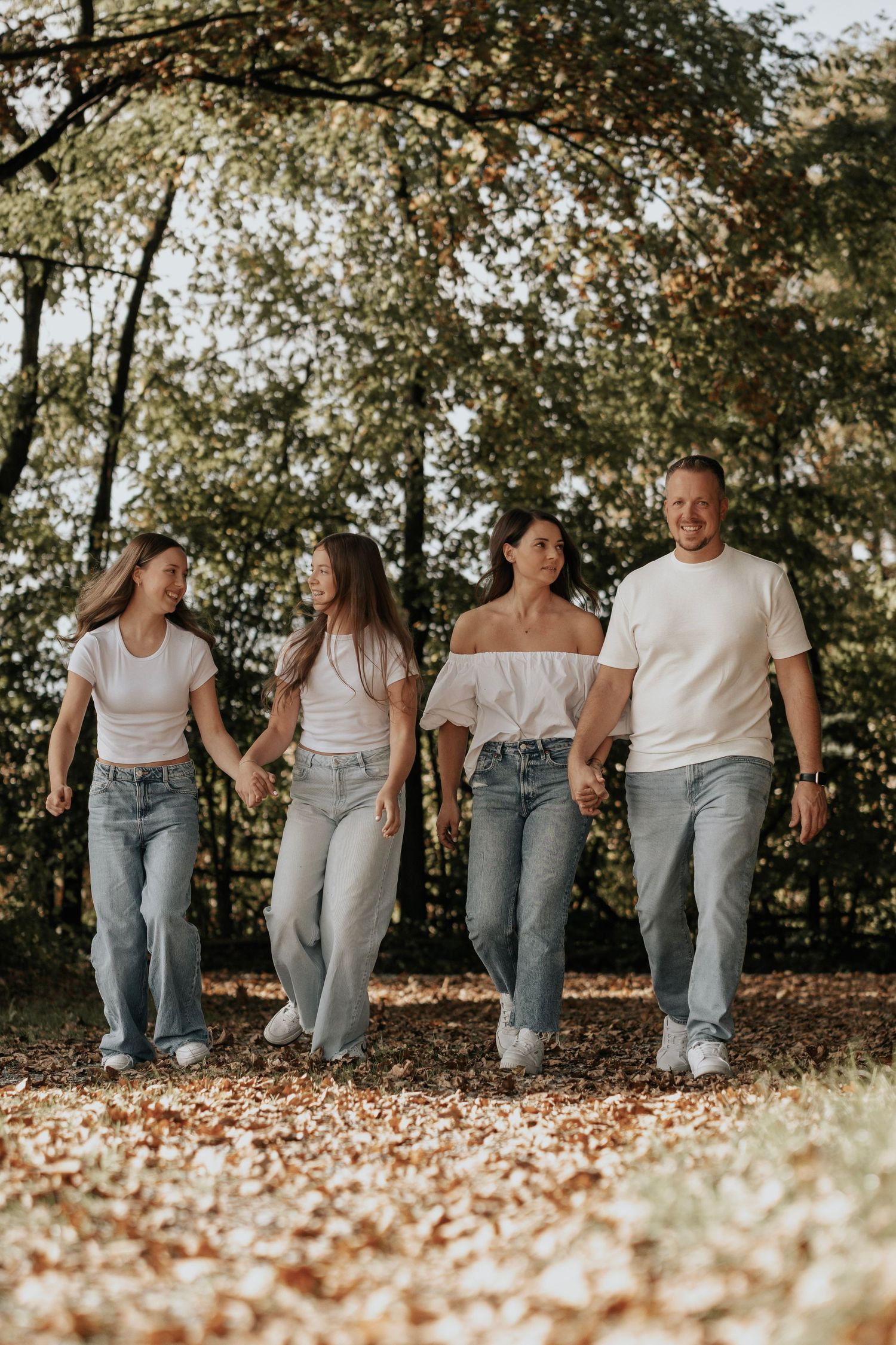 Happy family of four in matching white tops and jeans walking together in an autumn park.
