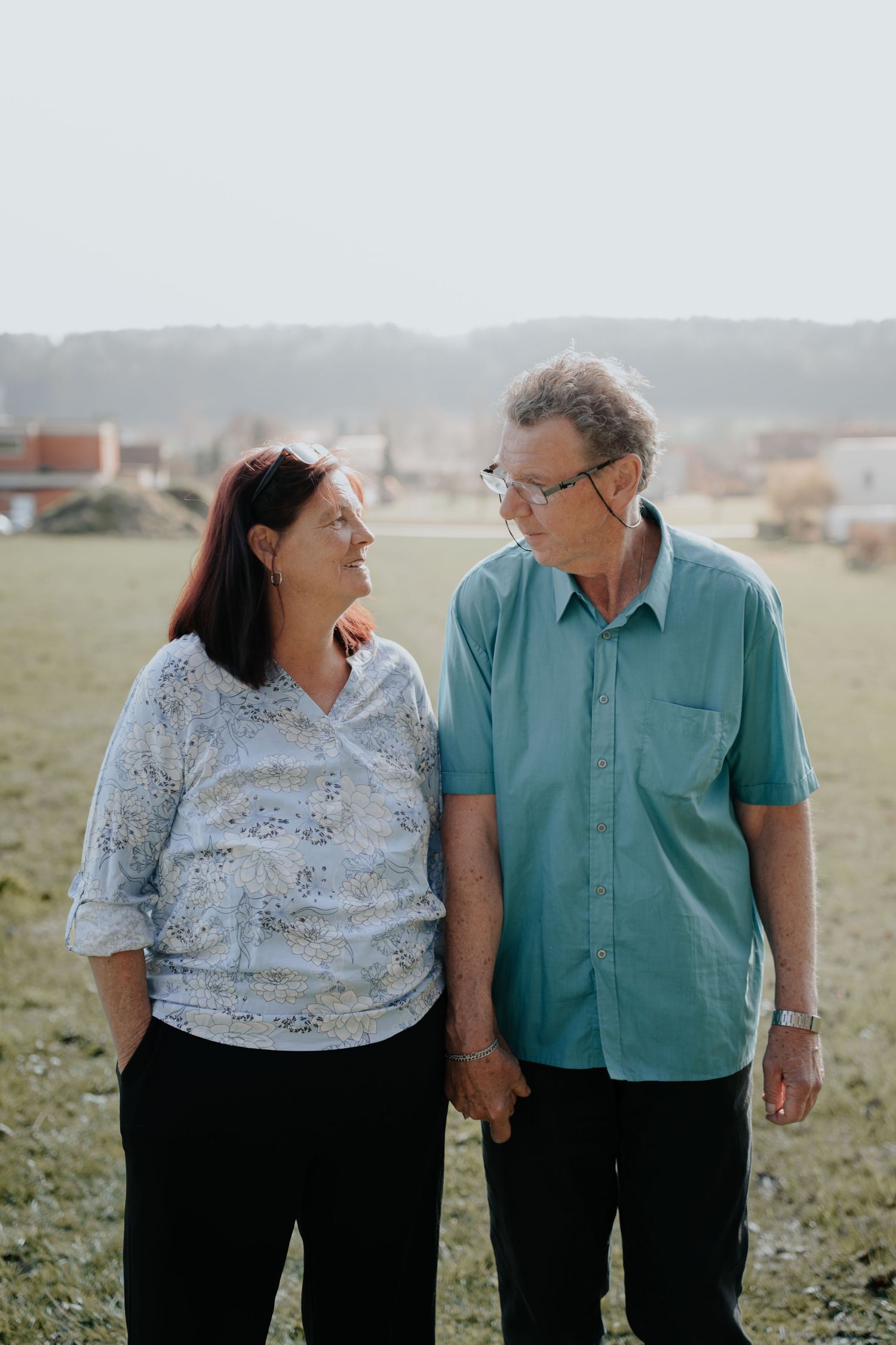 Middle-aged couple holding hands and smiling at each other in a sunny outdoor field setting.